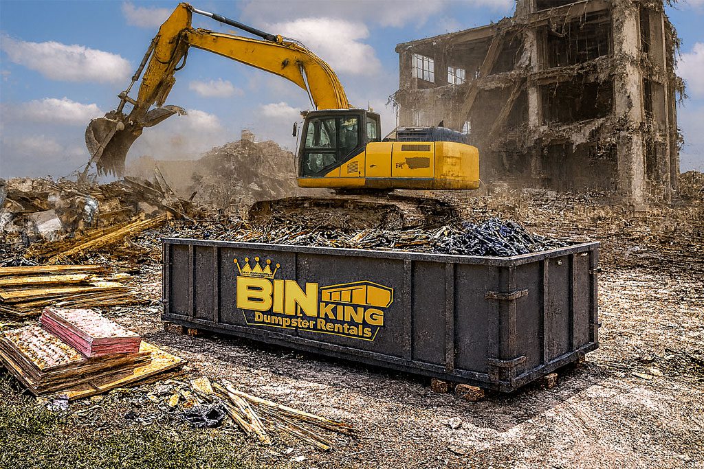 Bin King dumpster next to an excavator clearing concrete and building parts, used for site cleanup during renovations.